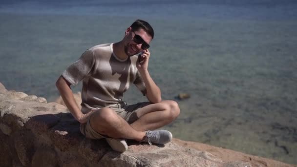 Adam kayalık Sahilde taş bankta oturur ve telefonda konuşmak. — Stok video