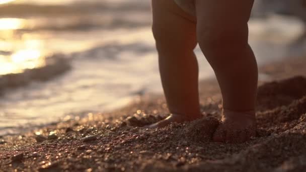 Increíbles piernas del bebé que hace pasos en la playa de arena al atardecer . — Vídeos de Stock