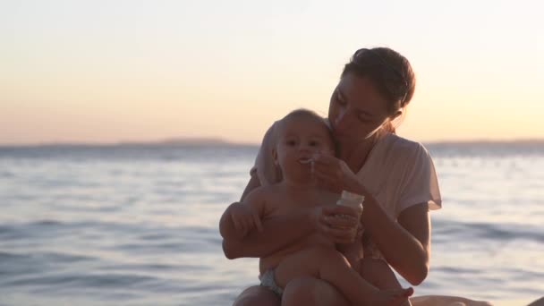 Mama karmi dziecko łyżeczką z słoika na plaży w pobliżu oceanu. — Wideo stockowe