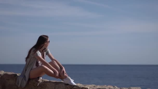 Kız taş çit üzerinde oturur ve deniz güzel bir görünüme sahiptir. — Stok video