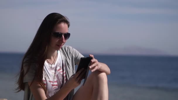 Een jong meisje zit op het strand in een zonnebril en maakt een telefoontje. — Stockvideo