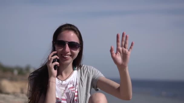 Дівчина сидить на пляжі в сонцезахисні окуляри і робить телефонний дзвінок. — стокове відео