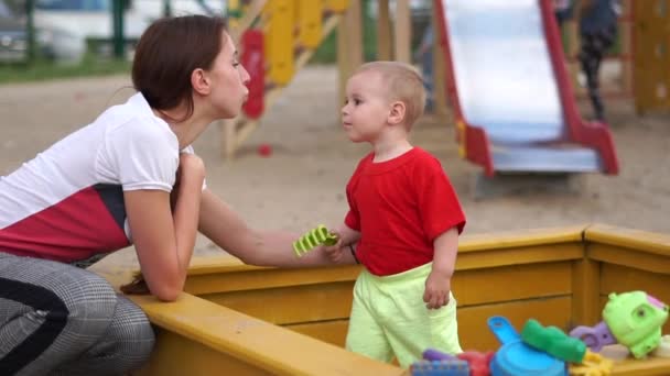 Молодая мать целует маленького мальчика в щеку на детской площадке в замедленной съемке — стоковое видео