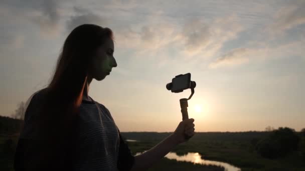 Όμορφο κορίτσι πυροβολεί βίντεο στο τηλέφωνο με έναν σταθεροποιητή στο ηλιοβασίλεμα, αργή κίνηση — Αρχείο Βίντεο