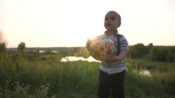 Sevimli bir küçük çocuk yavaş çekimde gün batımında çayırda bir top tutar — Stok video