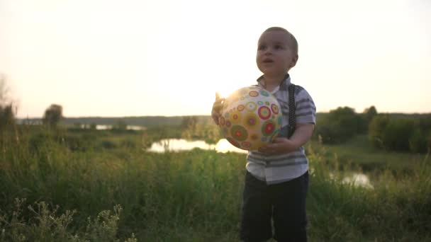 Roztomilá chlapeček drží míč na louce při západu slunce v pomalém pohybu — Stock video