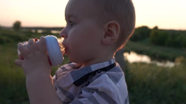 Uma criança bebe uma compota de uma mamadeira ao ar livre ao pôr do sol em câmera lenta — Vídeo de Stock