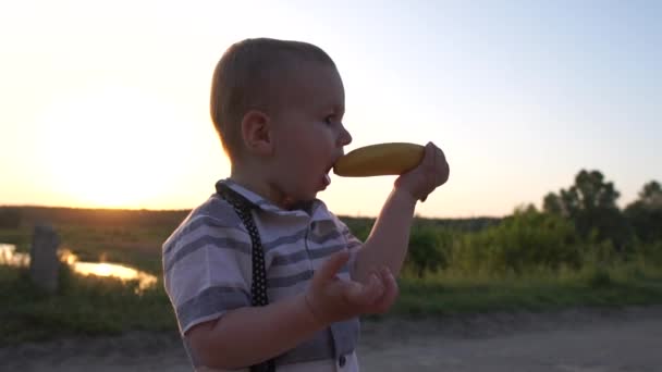Un garçon mignon prend une banane non pelée dans sa bouche dans le champ, au ralenti — Video