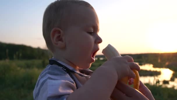 Aranyos kis fiú étkezési banán a parkban, és boldogan fut napnyugtakor, lassított — Stock videók