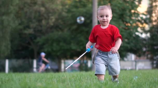 Um bebê bonito em shorts jeans correndo atrás de bolhas de sabão em câmera lenta ao ar livre — Vídeo de Stock