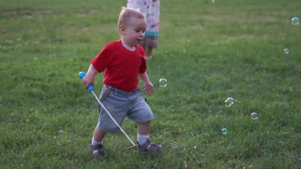 A little blond boy runs and catches soap bubbles in slow motion — Stock Video