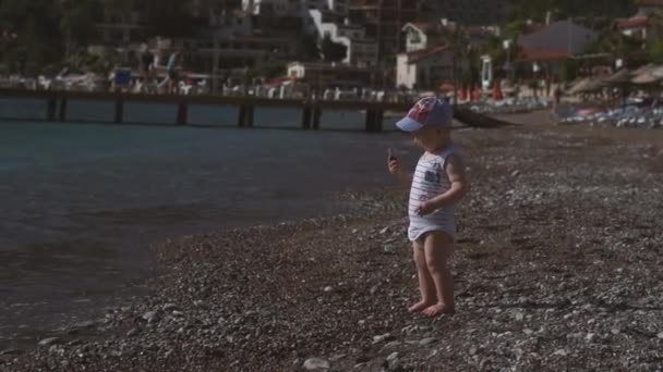 Милый малыш стоит на берегу моря, бросает камешек в воду, замедленное движение — стоковое видео