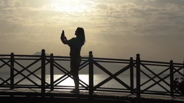 Uma jovem faz uma foto em um smartphone, de pé na ponte ao nascer do sol — Vídeo de Stock