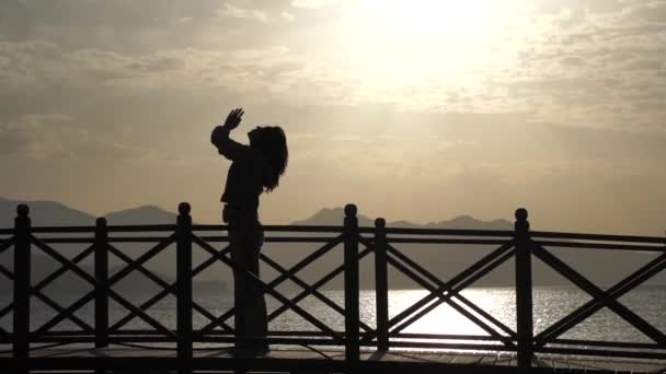Женщина, занимающаяся йогой на мосту против моря на закате — стоковое видео