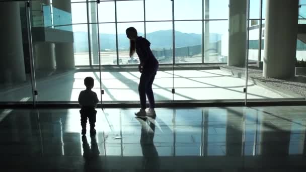 Мама и маленький мальчик идут к окну в зале аэропорта, чтобы посмотреть на самолет — стоковое видео