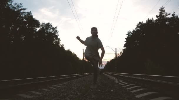 Un tipo en vaqueros rasgados y camiseta bailando krump entre la vía del tren contra el sol, cámara lenta — Vídeos de Stock