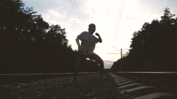 Un tipo con jeans rasgados y camiseta bailando hip hop entre las vías del tren contra el sol — Vídeos de Stock