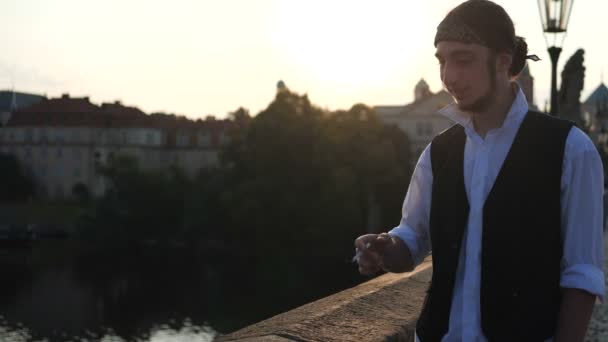 4k - Un niño sonriendo fumando en un puente cerca de los edificios antiguos al atardecer — Vídeos de Stock