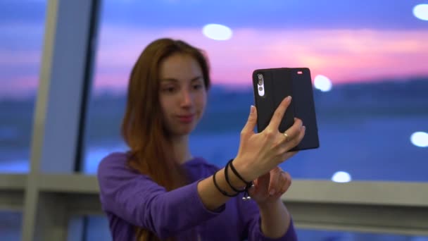 Mladá dívka dělá na telefonu v blízkosti okna na letišti selkou fotografii — Stock video