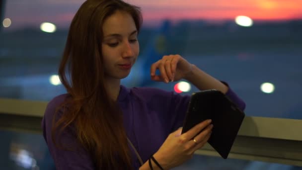 A student girl uses a smartphone, standing at the airport waiting for her flight — Stock Video