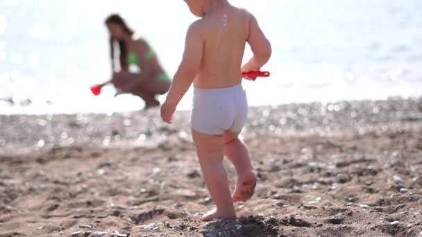 En liten bebis går längs stranden och bär en spade till havet i slow motion — Stockvideo
