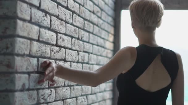 Chica rubia inspirada caminando a lo largo de una pared de ladrillo y tocarlo en un estudio en slo-mo — Vídeo de stock