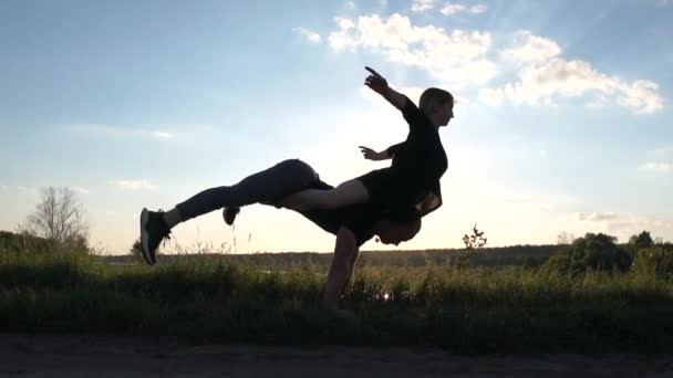 Brave uomo biondo fa un handstand con una ragazza sulla schiena a un lago al tramonto in slo-mo — Video Stock