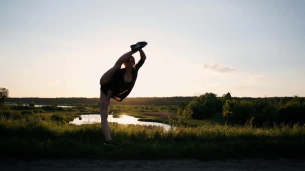 Cheerful girl standing and doing a straddle oversplit at a pond at sunset in slo-mo — Stock Video