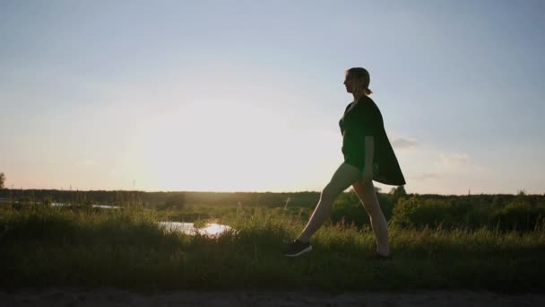 Sportliche junge Frau, die bei Sonnenuntergang im Slo-mo an einem See steht und einen Frontsplit macht — Stockvideo