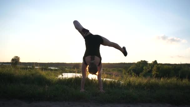 Rozciągliwa dziewczyna robi Handstand z pochyleniem dodatnim podzielone na jeziorze o zachodzie słońca w SLO-mo — Wideo stockowe