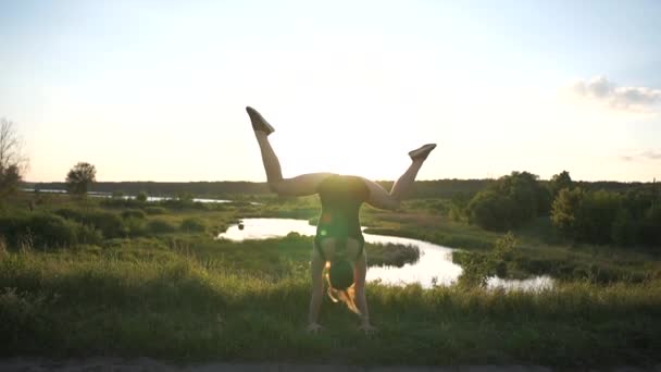 Wesoły dziewczyna robi Handstand z skrzyżowanymi nogami w stawie o zachodzie słońca w SLO-mo — Wideo stockowe