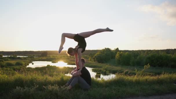 Hombre alegre se sienta y mantiene a su chica de rodillas haciendo una división frontal al atardecer en slo-mo — Vídeo de stock