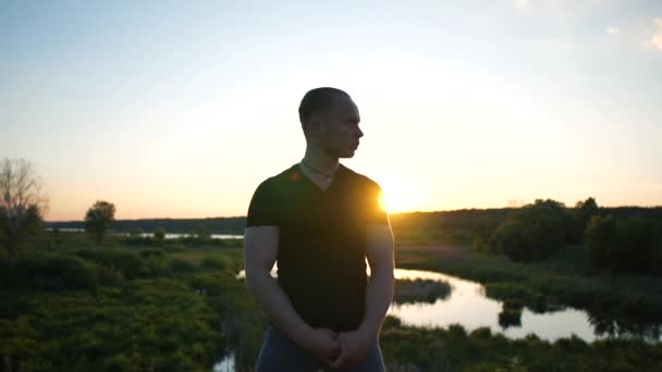 Modig man står i en grenslar sätt vrida huvudet åt sidan vid sjön vid solnedgången i slo-mo — Stockvideo