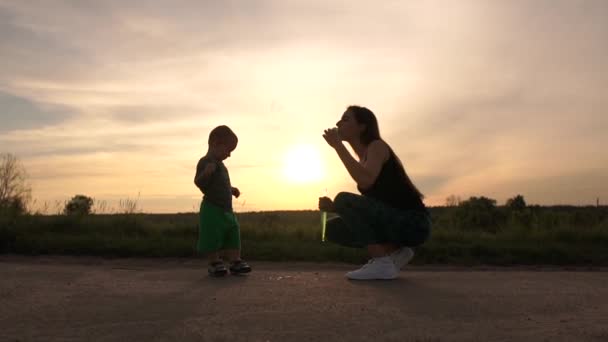Щаслива мати сидить і робить мильні бульбашки для своєї дитини на заході сонця в шлю-мо — стокове відео