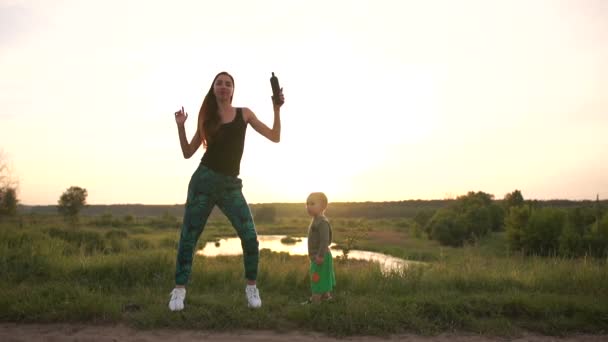 Gelukkige moeder dansen met haar peuter bij een klein meer bij zonsondergang in slo-mo — Stockvideo