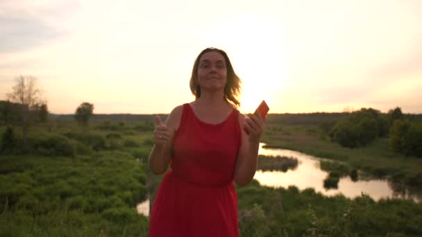 Уморительная женщина танцует со своим телефоном у маленького пруда с тростью на закате в тихом месте — стоковое видео