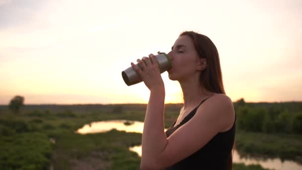Schlanke junge Frau trinkt Tee aus einer Flasche am Teich bei Sonnenuntergang in slo-mo — Stockvideo