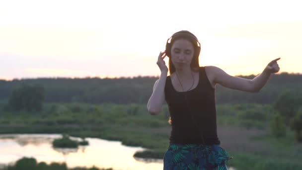 Chica feliz bailando en auriculares con una sensación en el lago al atardecer en slo-mo — Vídeo de stock