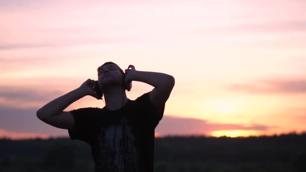 Hombre inspirado de pie en los auriculares y bailando en el lago al atardecer en slo-mo — Vídeos de Stock