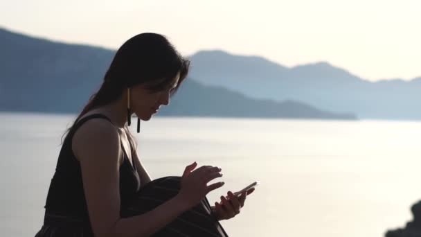 A girl types a message on the smartphone sitting against the sea in slow motion — Stock Video