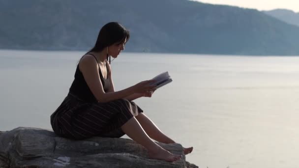 Una joven está leyendo un libro en un hermoso lugar contra el mar, en cámara lenta — Vídeo de stock