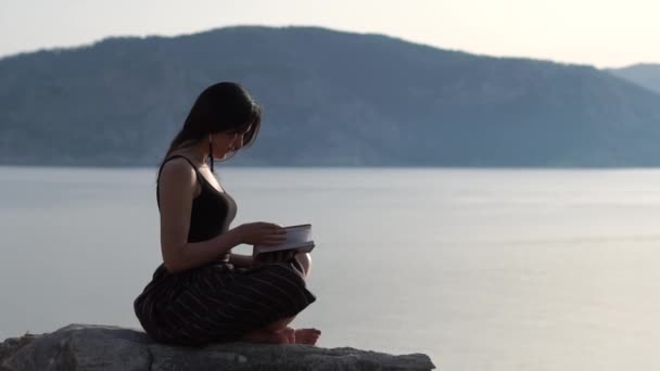 Ragazza romantica legge con entusiasmo un libro vicino al mare all'alba, al rallentatore — Video Stock