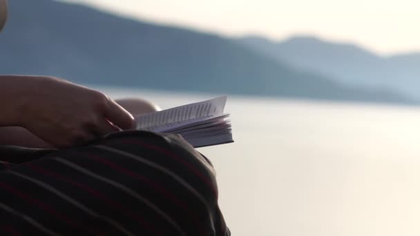 Kvinnan läser en bok nära havet, händer nära i slow motion vid soluppgången — Stockvideo