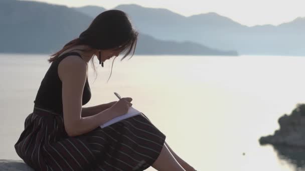 Mladá dívka píše v poznámkovém bloku a v pomalém pohybu sedí na pobřeží — Stock video