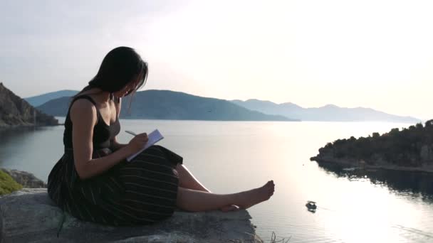 Una donna scrive in un taccuino seduta su una roccia sopra il mare al rallentatore — Video Stock