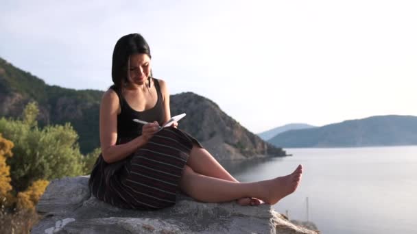 Seksi bir kız denizin üzerindeki bir kayanın üzerinde yalınayak oturan bir not defterine yazıyor. — Stok video