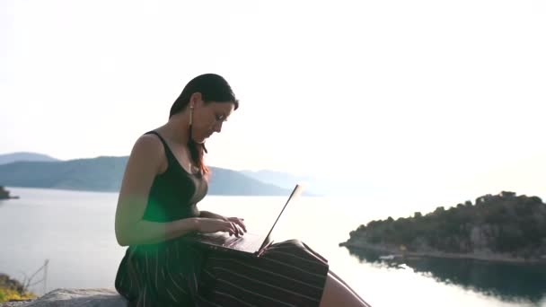 Ein junges Mädchen arbeitet an einem Laptop, sitzt auf einem Felsen über dem Meer, Zeitlupe — Stockvideo