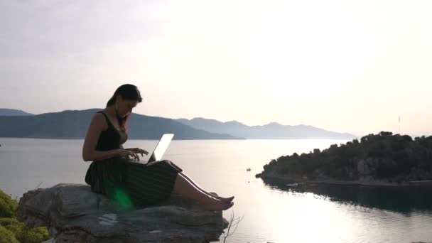 A pretty girl student works at a laptop on a rock above the sea, slow motion — Stock Video