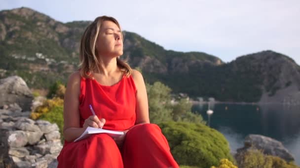 A woman makes notes in a notebook sitting on a rock above the sea in slow motion — Stock Video