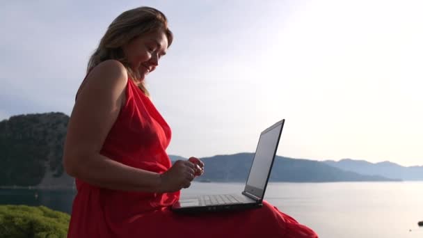 Улыбающаяся женщина, печатающая на ноутбуке, сидя против красивого морского пейзажа, замедленной съемки — стоковое видео